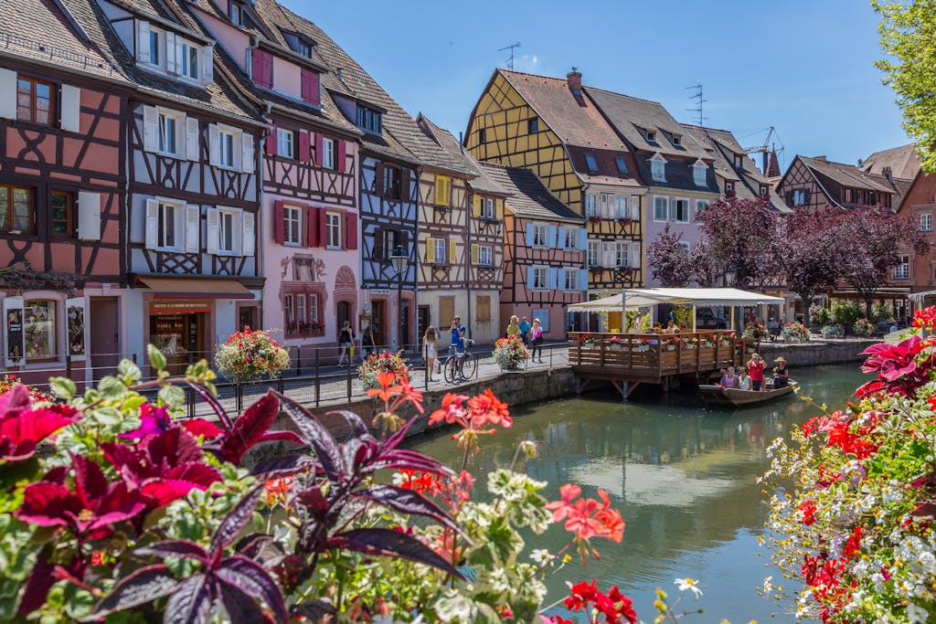 Colorful medieval houses along a canal in Colmar, France, with vibrant flowers and sunny skies.